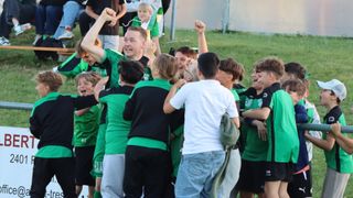 Eine Gruppe junger Fußballer in grünen Trikots feiert einen Sieg auf einem Rasenfeld. Ein Junge in einem weißen Hemd steht in der Mitte, umgeben von jubelnden Teamkollegen.