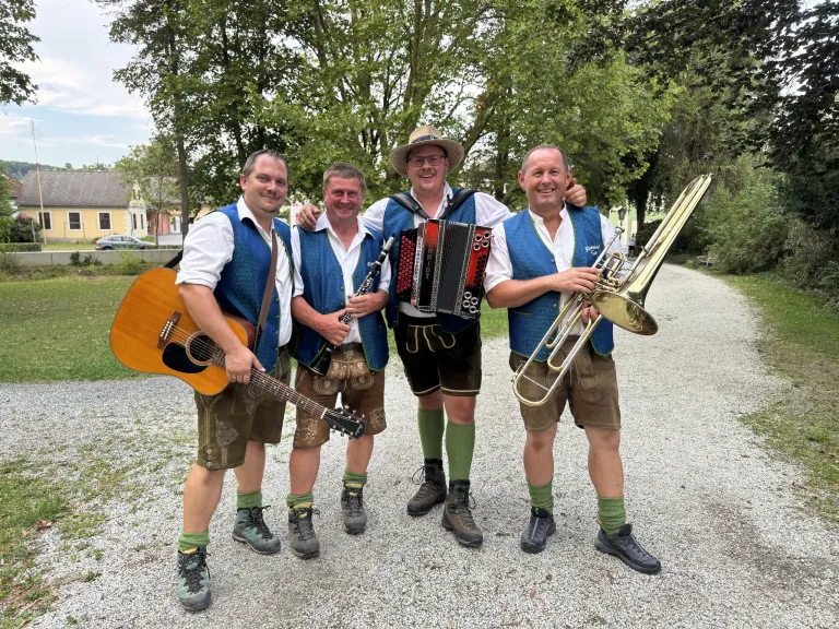 Vier Männer in traditioneller bayerischer Kleidung posieren mit Musikinstrumenten für ein Foto. Von links nach rechts hält ein Mann eine Gitarre, ein anderer eine Klarinette, ein dritter ein Akkordeon und der letzte eine Trompete. Sie stehen auf einem Kiesweg mit Bäumen und einem Haus im Hintergrund.
