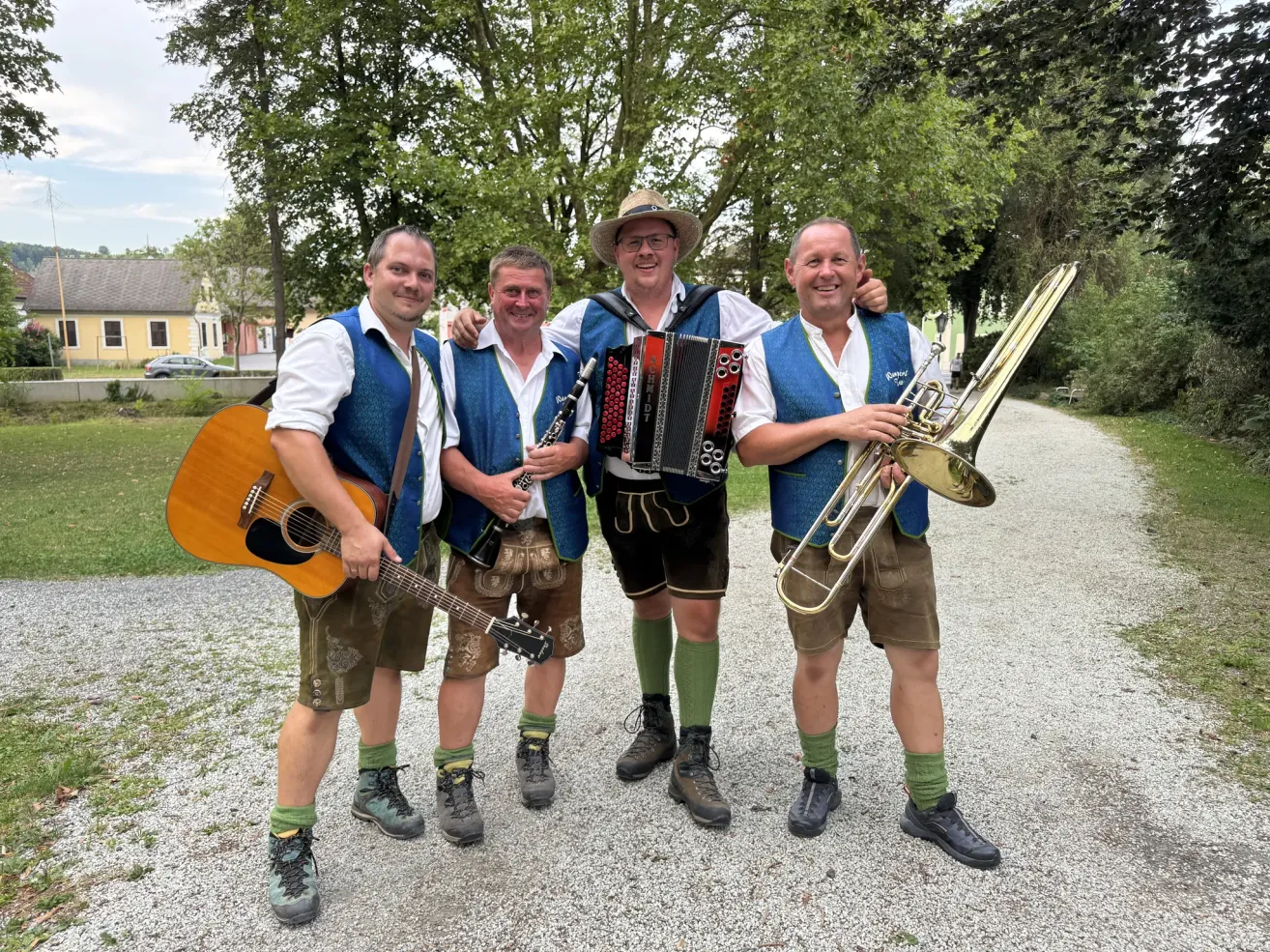 Vier Männer in traditioneller bayerischer Kleidung posieren mit Musikinstrumenten für ein Foto. Von links nach rechts hält ein Mann eine Gitarre, ein anderer eine Klarinette, ein dritter ein Akkordeon und der letzte eine Trompete. Sie stehen auf einem Kiesweg mit Bäumen und einem Haus im Hintergrund.