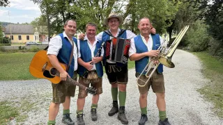 Vier Männer in traditioneller bayerischer Kleidung posieren mit Musikinstrumenten für ein Foto. Von links nach rechts hält ein Mann eine Gitarre, ein anderer eine Klarinette, ein dritter ein Akkordeon und der letzte eine Trompete. Sie stehen auf einem Kiesweg mit Bäumen und einem Haus im Hintergrund.
