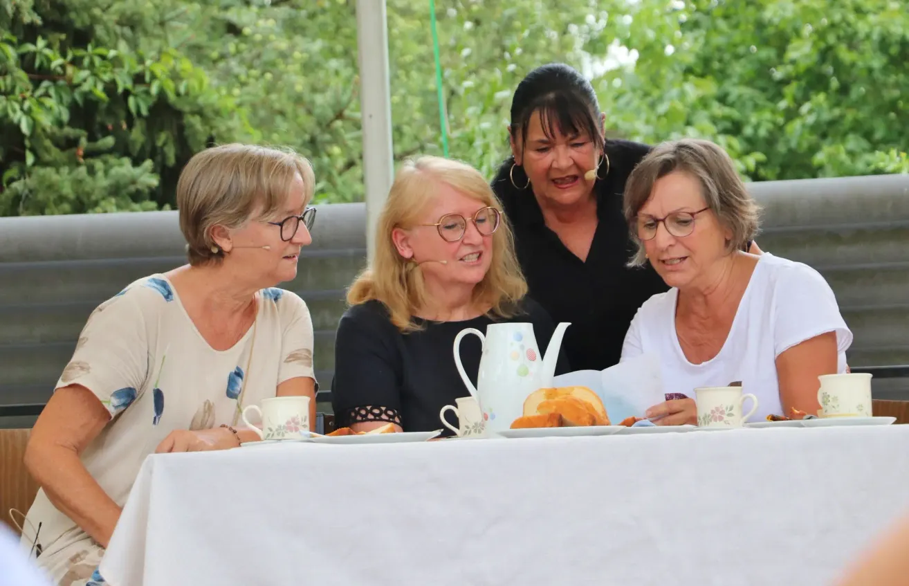 Vier Frauen sitzen an einem Tisch, lächeln und unterhalten sich. Eine Frau hält ein Mikrofon. Hinter ihnen steht eine Frau und spricht in ein Mikrofon.