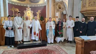 Eine Gruppe von Priestern steht in einer Kirche zusammen, einer mit einem Stock, ein anderer hält ein Buch, und eine Statue eines Heiligen hinter ihnen.