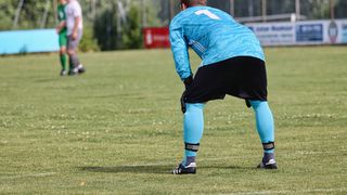 Ein Fußballspieler in einem blauen Trikot mit der Nummer 1 auf dem Rücken, beugt sich auf einem Rasenfeld. Im Hintergrund stehen zwei Spieler in grünen und weißen Uniformen.