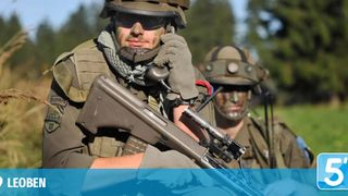 Zwei Soldaten in Tarnuniformen und Helmen halten ein Gewehr, einer schaut in die Kamera.