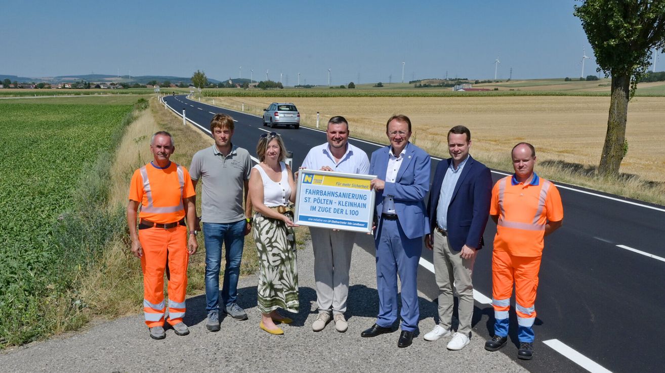 Eine Gruppe von Menschen, darunter zwei Frauen, steht auf einer Straße mit einem Schild, das 'Fahrbahnsanierung St. Polten - Kleinhain im Zuge der L100' besagt. Sie sind in verschiedene Kleidung gekleidet, einige tragen Sicherheitswesten. Die Straße hat eine neue Asphaltoberfläche, und im Hintergrund gibt es ein Feld und ein Auto auf der Straße.