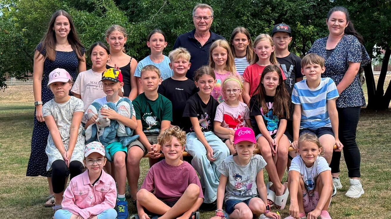 Eine Gruppe von Kindern und ein erwachsener Mann posieren für ein Foto im Freien. Die Kinder sitzen auf einer Bank und lächeln, während der Mann hinter ihnen steht.