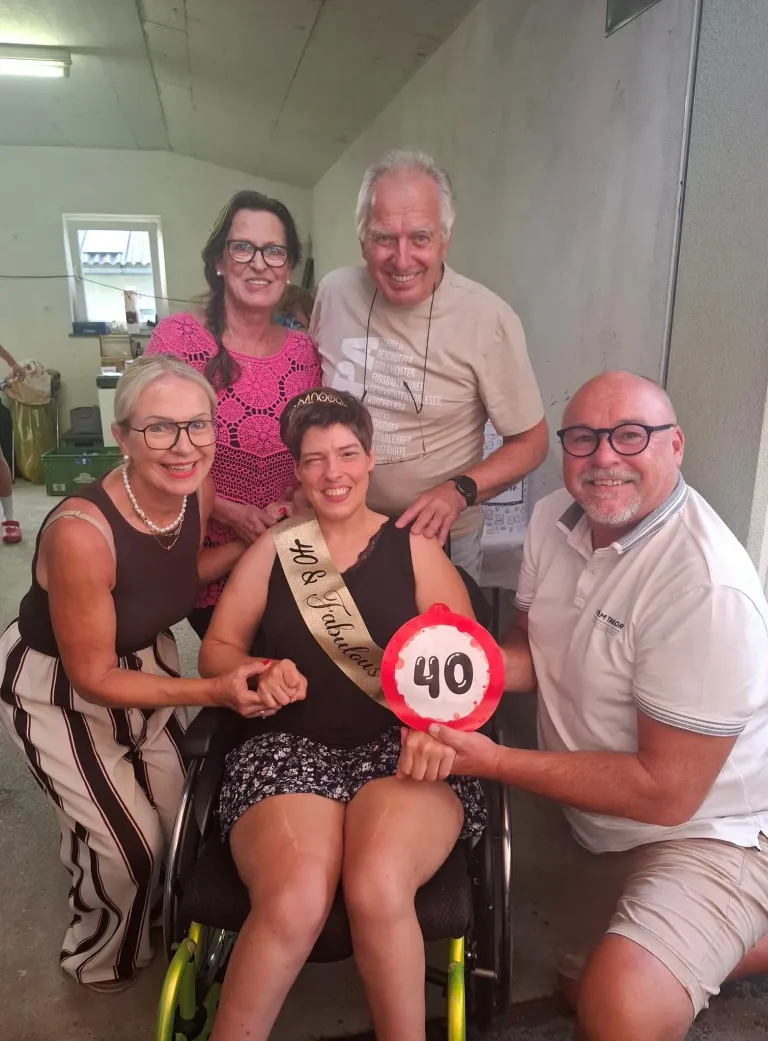 Fünf Personen posieren für ein Foto, wobei eine Frau im Rollstuhl eine Schärpe mit der Aufschrift '40 Fabulous' trägt. Die Frau im Rollstuhl lächelt und hält ein Schild mit der Nummer 40.