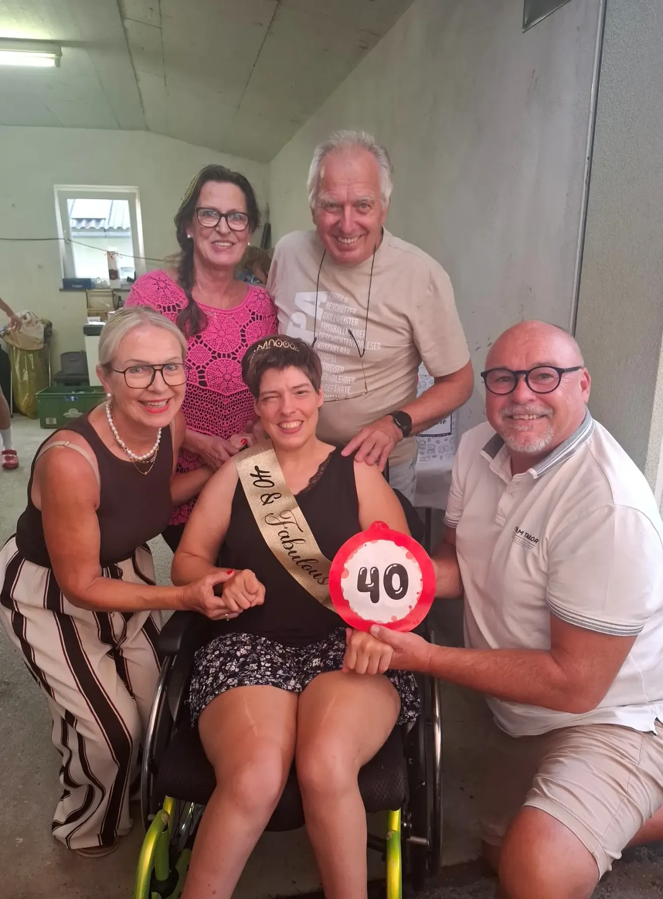 Fünf Personen posieren für ein Foto, wobei eine Frau im Rollstuhl eine Schärpe mit der Aufschrift '40 Fabulous' trägt. Die Frau im Rollstuhl lächelt und hält ein Schild mit der Nummer 40.
