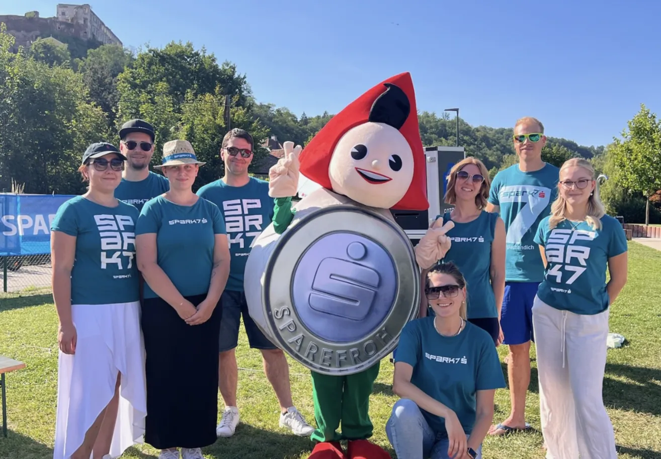 Eine Gruppe von Menschen in passenden blauen T-Shirts posiert mit einem Maskottchen, das ein großes kreisförmiges Objekt mit der Aufschrift 'SPAREFF' hält. Sie stehen auf einem Rasenfeld mit Bäumen und einem klaren Himmel im Hintergrund.