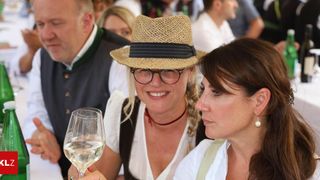 Zwei Frauen bei einem gesellschaftlichen Treffen, eine mit Strohhut und Weinglas, die andere mit geflochtenen Haaren und Ohrringen.