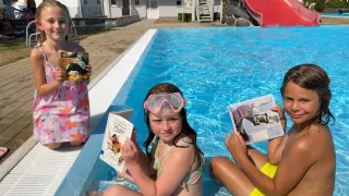 Drei Kinder sitzen in einem Pool, zwei von ihnen lesen Bücher, während das dritte ein Buch in der Hand hält. Sie tragen alle Badekleidung. Hinter ihnen steht ein Gebäude mit einer roten Rutsche.