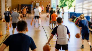 Eine Gruppe von Kindern spielt Basketball in einer Turnhalle. Einige dribbeln den Ball, während andere in einer Reihe stehen. Es gibt orangefarbene Kegel auf dem Boden.