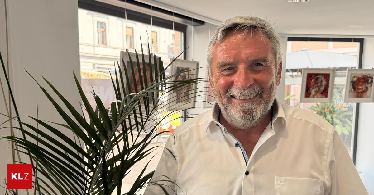 Ein Mann mit weißem Bart und Schnurrbart lächelt vor einem Fenster mit einer Pflanze und gerahmten Bildern im Hintergrund.