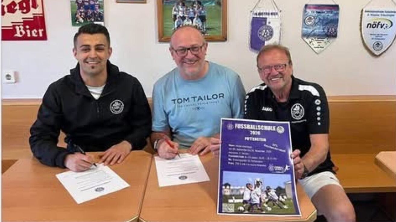 Drei Männer sitzen an einem Tisch, lächeln und halten Stifte. Ein Mann unterschreibt ein Dokument, während ein anderer ein Plakat für eine Fußballschule hält.