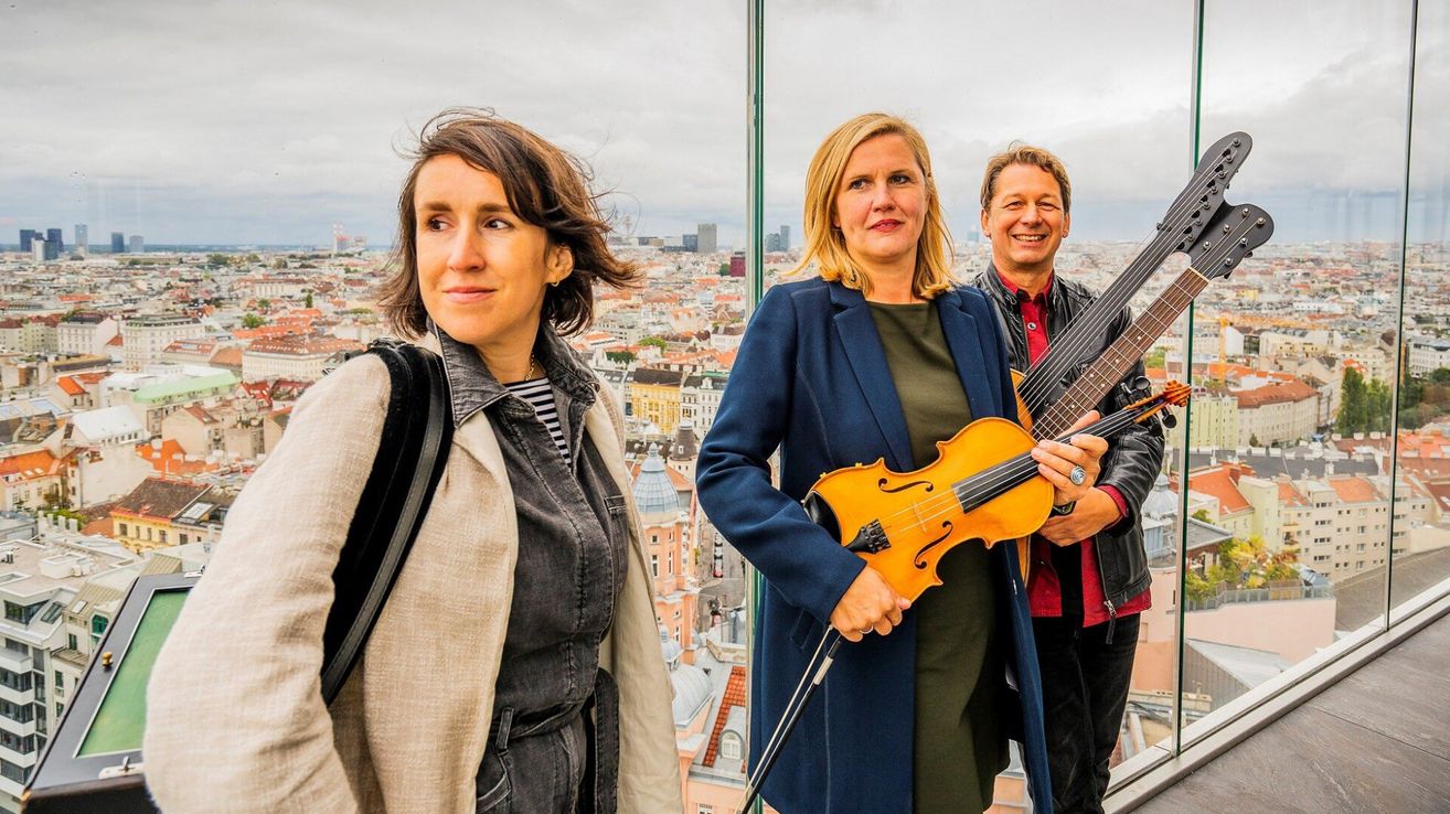 Drei Musiker stehen auf einem Balkon mit Blick auf die Stadt. Eine Frau in einem blauen Mantel hält eine Geige, während ein Mann in einem schwarzen Lederjacken eine Gitarre hält. Eine weitere Frau in einem beigen Mantel lächelt.