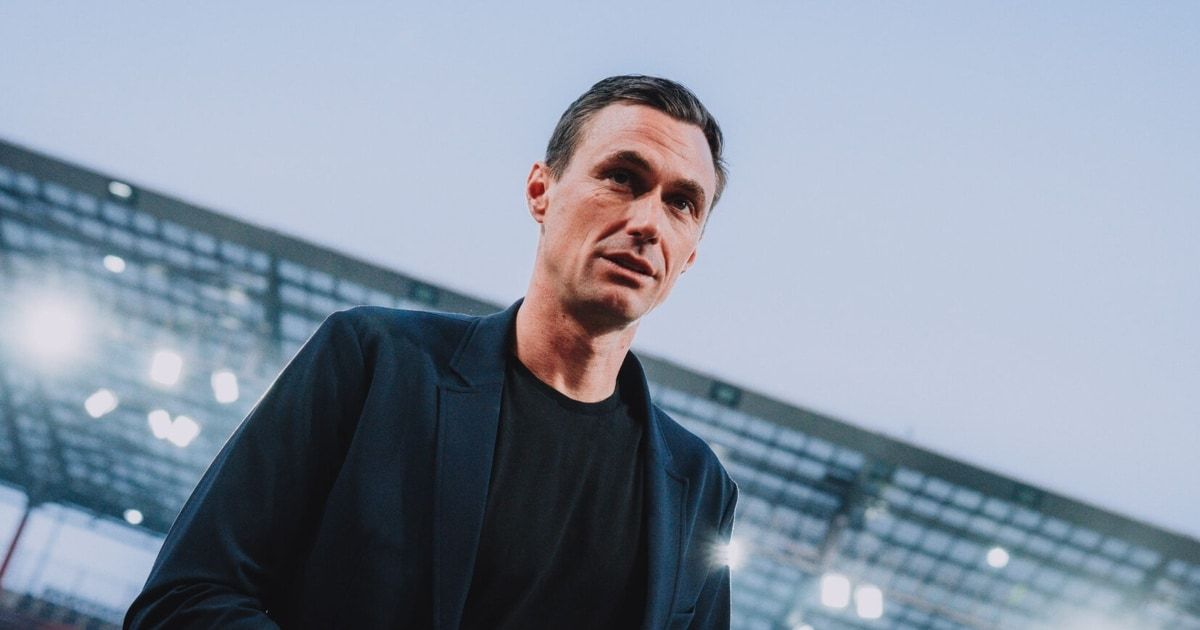 Ein Mann in einem schwarzen Shirt und einer Jacke steht vor einem Stadion und sieht ernst aus. Das Stadion ist groß mit einem modernen Design, und der Himmel ist klar.