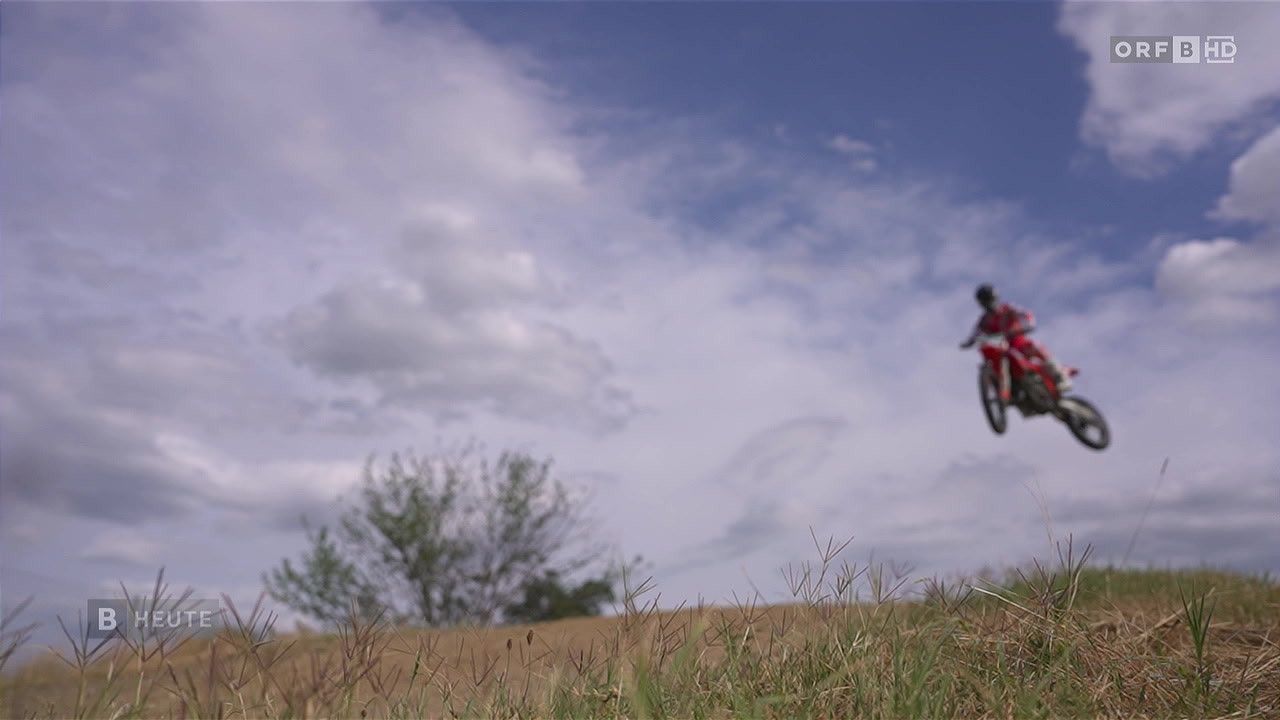 Eine Person fährt ein rotes Motorrad in der Luft, möglicherweise führt sie einen Stunt durch. Der Himmel ist blau mit verstreuten Wolken, und im Hintergrund befindet sich ein kleiner Baum.