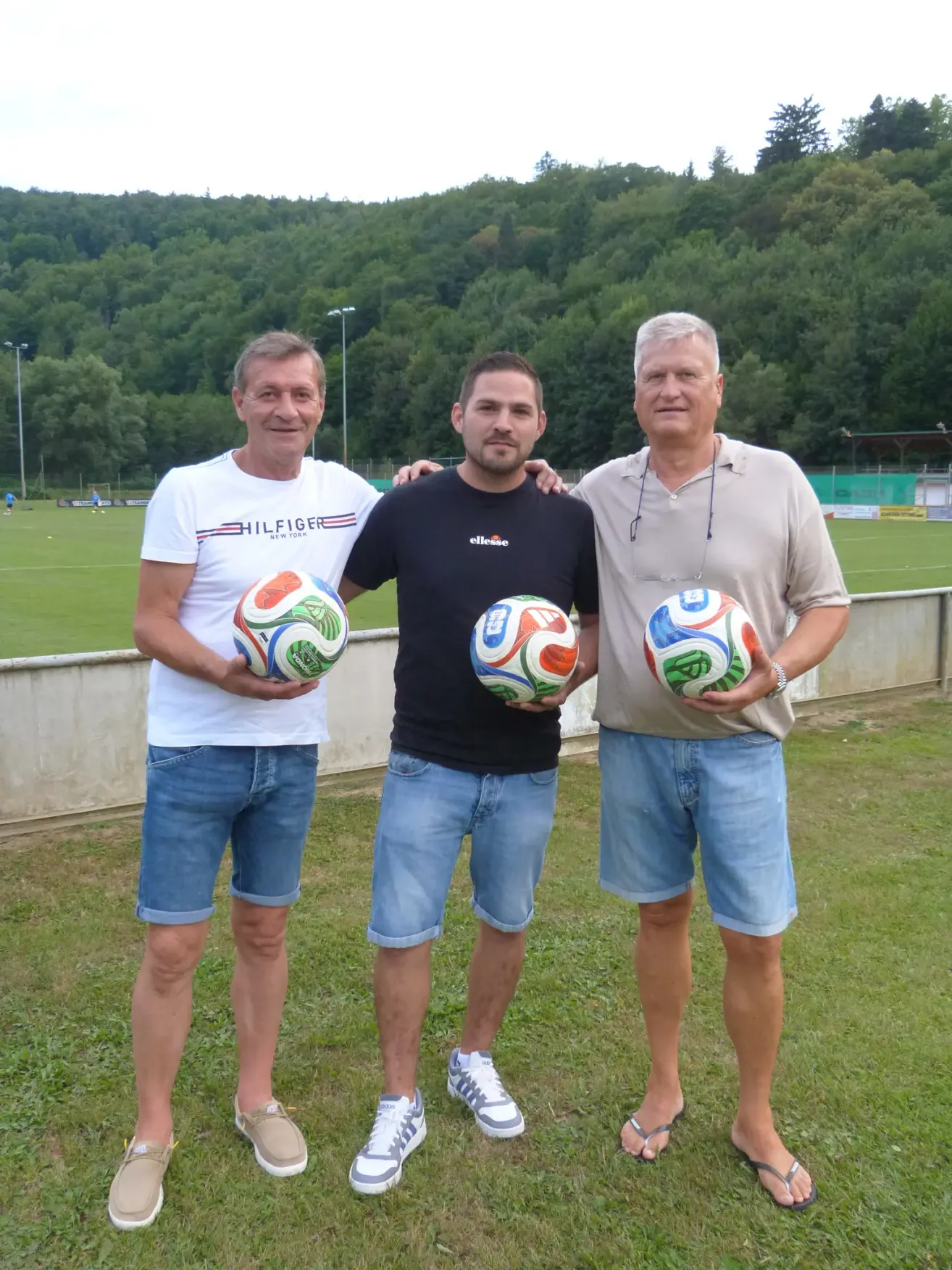 Drei Männer stehen auf einem Rasenfeld, jeder hält einen Fußball. Sie sind lässig gekleidet und scheinen für ein Foto zu posieren. Dahinter befindet sich ein Rasenbereich mit einer Betonbarriere und einem Fußballfeld.