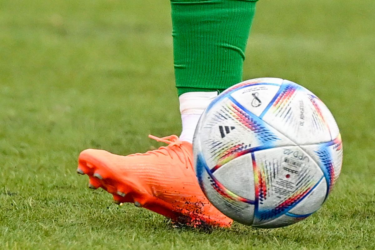 Ein Fußballspieler in grünen Socken und orangefarbenen Stollenschuhen tritt einen Fußball mit einem farbenfrohen Design.