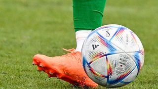 Ein Fußballspieler in grünen Socken und orangefarbenen Stollenschuhen tritt einen Fußball mit einem farbenfrohen Design.