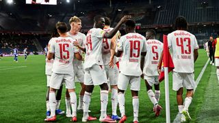 Eine Fußballmannschaft in weißen Trikots mit Red Bull-Branding feiert auf dem Feld bei Nacht. Spieler mit den Nummern 15, 19 und 43 sind sichtbar.