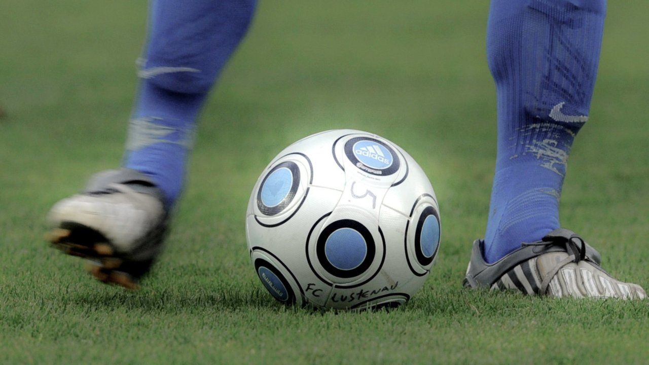 Ein Fußball mit blauem und schwarzem Design, von Adidas gebrandet, liegt auf einem grünen Feld mit der blauen Uniform eines Spielers im Hintergrund.