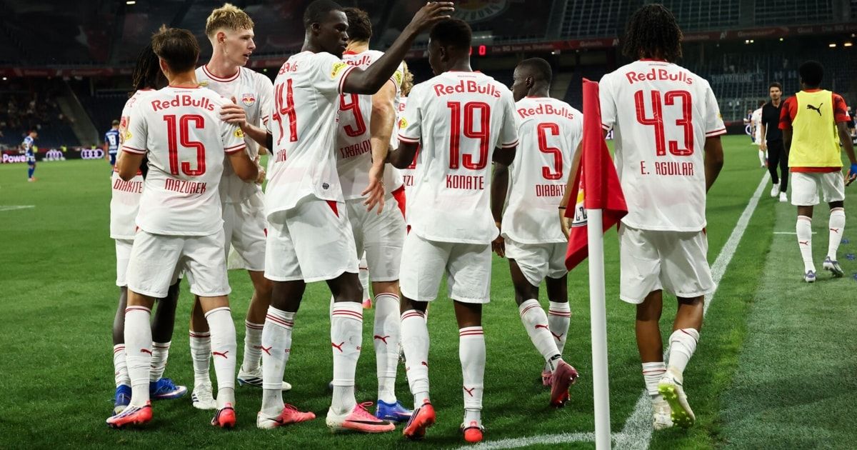 Eine Gruppe von Fußballspielern in weißen Trikots mit roten Akzenten steht auf dem Spielfeld. Sie tragen Turnschuhe und haben Nummern auf dem Rücken.