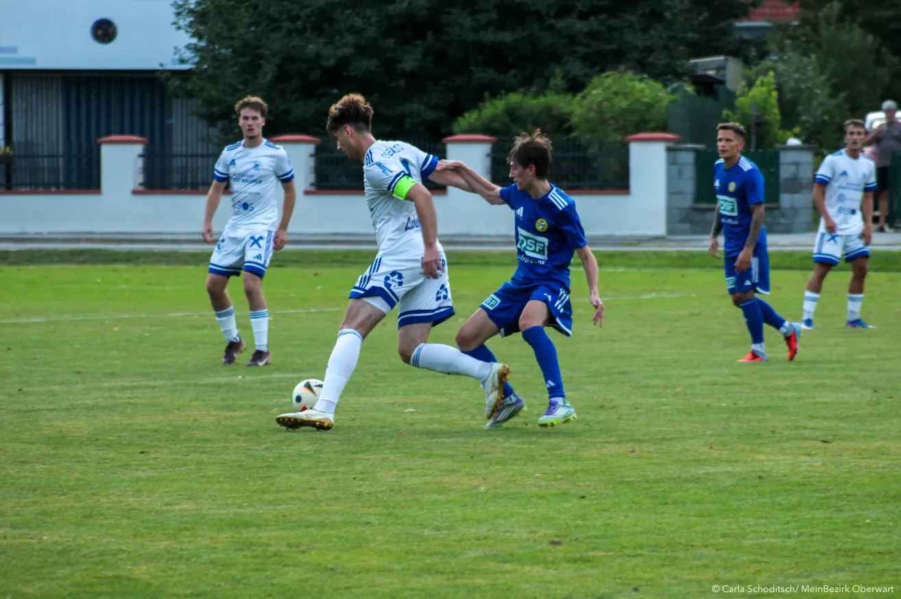 Zwei Fußballspieler in blauen und weißen Trikots kämpfen auf einem Rasenfeld um den Ball. Ein weiterer Spieler in Weiß beobachtet von hinten.