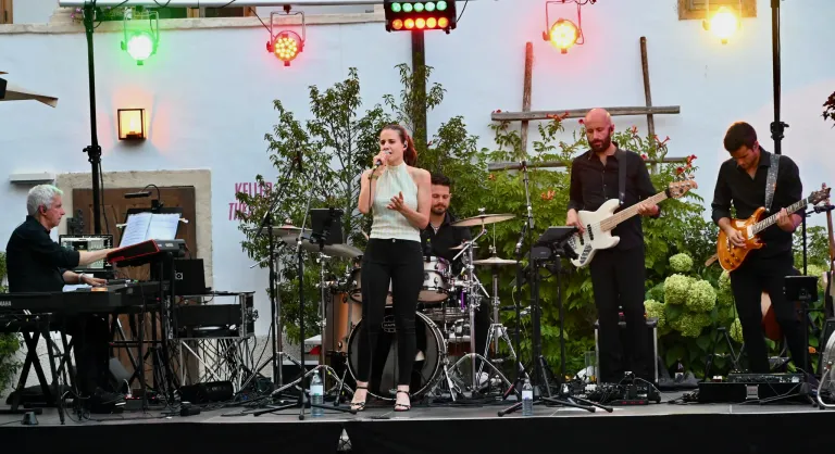 Eine Frau singt auf der Bühne mit einer Band, bestehend aus einem Schlagzeuger und einem Gitarristen, unter Bühnenbeleuchtung und vor Grünpflanzen.