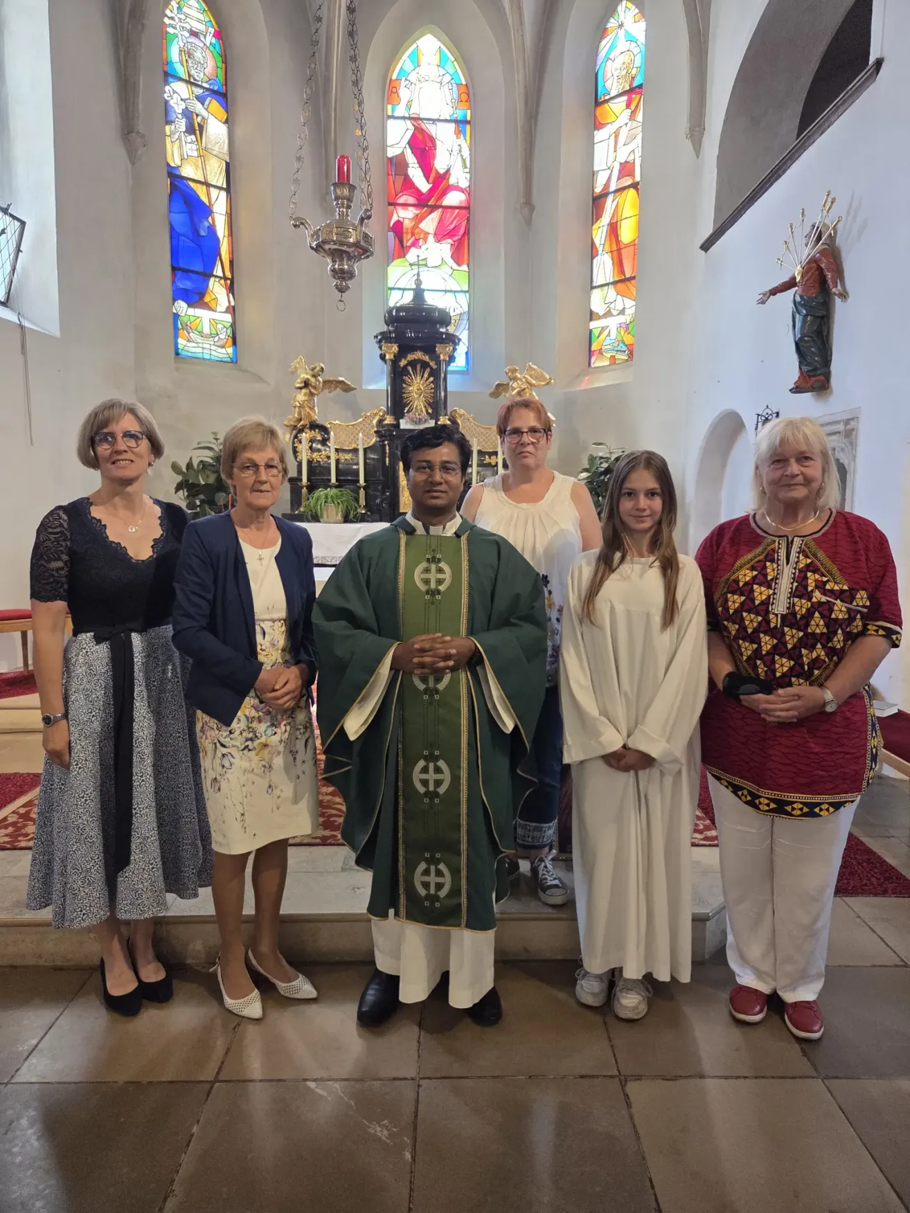 Eine Gruppe von Menschen, einschließlich eines Priesters, steht vor einem Kirchenaltar. Der Priester trägt eine grün-weiße Robe, die anderen tragen Freizeitkleidung. Dahinter steht eine Statue und es gibt Buntglasfenster.