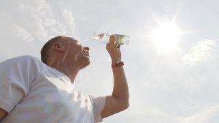Ein Mann trinkt Wasser aus einer Flasche im Freien unter einem hellen Himmel mit Wolken.