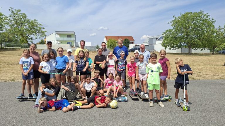 Eine Gruppe von Kindern und Erwachsenen posiert für ein Foto im Freien, einige mit Skateboards und andere mit Sportgeräten. Im Hintergrund sind Häuser und Bäume zu sehen.