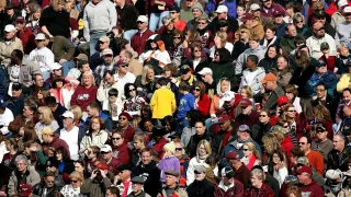 Eine große Menschenmenge, einige mit Mützen und Brillen, versammelt sich in einem Stadion. Viele tragen Kleidung in den Farben Rot und Weiß.