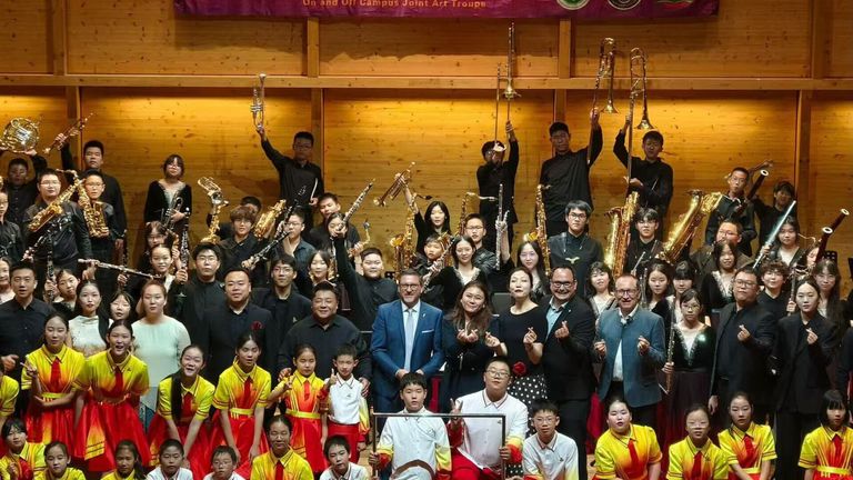 Eine Gruppe von Musikern, einschließlich Kinder, Erwachsene und Lehrer, posiert für ein Foto mit ihren Instrumenten. Sie sind in passenden Uniformen gekleidet und stehen vor einer Holzwand.