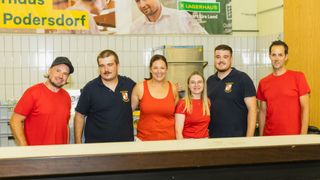 Vier Personen stehen hinter einem Tresen und lächeln für ein Foto. Zwei Männer und zwei Frauen, alle tragen schwarze Shirts mit Logos. Eine Frau in einem orangefarbenen Tanktop steht zwischen den Männern. Dahinter ist eine geflieste Wand mit einer großen Werbung.