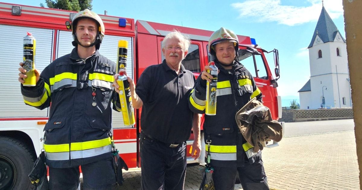 Drei Personen, zwei in Feuerwehruniformen und eine in einem schwarzen Hemd, stehen vor einem roten Feuerwehrauto. Die Feuerwehrleute halten gelbe Kanister, und der Mann im schwarzen Hemd lächelt.