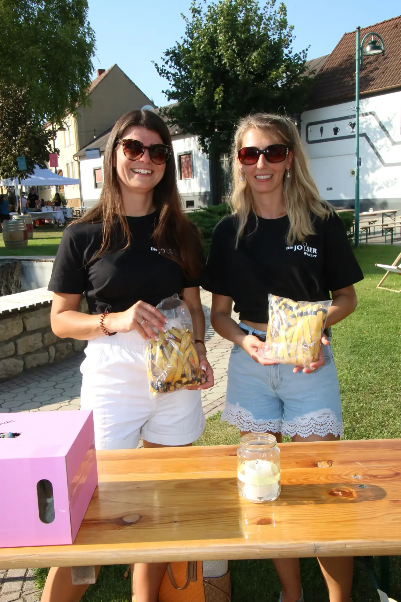 Zwei Frauen in passenden schwarzen T-Shirts stehen an einem Tisch im Freien, lächeln und halten Tüten mit gelben Snacks. Die Frau links trägt weiße Shorts und ein Armband, während die Frau rechts Jeansshorts mit Spitzenbesatz trägt. Ein rosafarbener Kasten und ein Glas mit einer Kerze stehen auf dem Tisch.