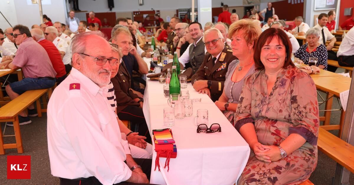 Eine Gruppe von Menschen, darunter ein Mann in Uniform, sitzt an einem langen Tisch mit verschiedenen Gegenständen darauf, wie Gläser, Flaschen und ein roter Stoff. Sie scheinen sich auf einer gesellschaftlichen Veranstaltung zu befinden.