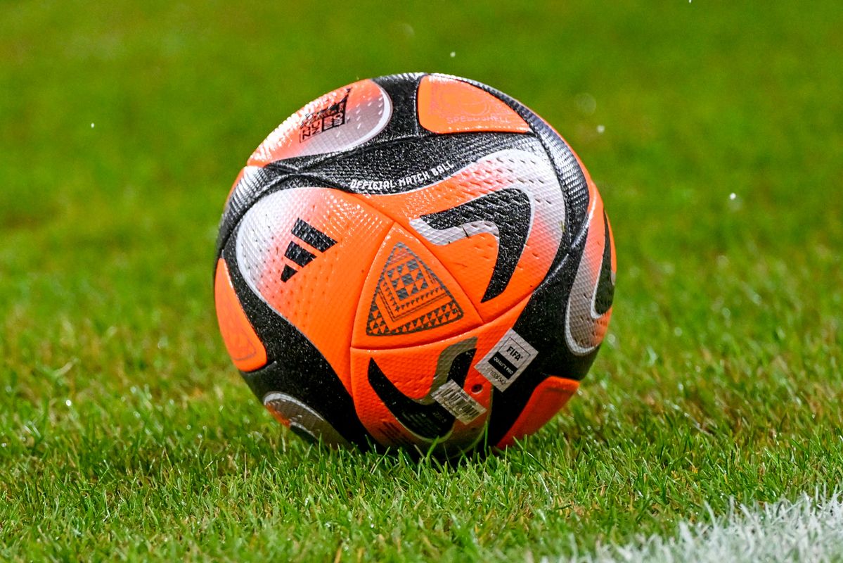 Nahaufnahme eines Fußballs mit orange, schwarz und weißen Farben, der auf einem Rasen liegt. Der Ball hat ein geometrisches Muster und einen FIFA-Qualitätsstempel.