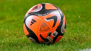 Nahaufnahme eines Fußballs mit orange, schwarz und weißen Farben, der auf einem Rasen liegt. Der Ball hat ein geometrisches Muster und einen FIFA-Qualitätsstempel.