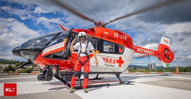 Eine Person in einem roten Overall und Helm steht neben einem roten Hubschrauber mit der Aufschrift 'ARA Flugrettung' an der Seite. Der Hubschrauber hat die Registrierungsnummer 'OE-XFR' und ist auf einem Rollfeld geparkt. Der Himmel ist bewölkt.
