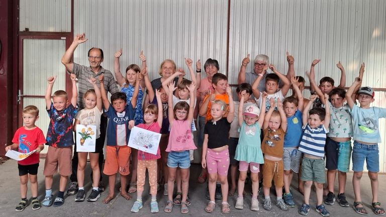 Eine Gruppe von Kindern und Erwachsenen steht zusammen, hebt die Hände und lächelt für ein Foto in einem Raum mit einer weißen Wand.