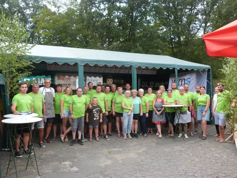 A group of people wearing green shirts are posing for a photo in front of a tent. There are trees in the background.