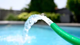 Ein grüner Schlauch sprüht Wasser in einen Pool, wobei das Sonnenlicht von den Wassertropfen reflektiert wird.