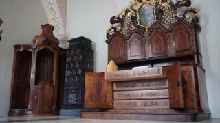 Ein kunstvoll verzierter hölzerner Schrank mit goldenen Akzenten, offenen Schubladen und einer dekorativen Malerei darüber. Dahinter befindet sich ein schwarzer, kunstvoll gestalteter Gegenstand.