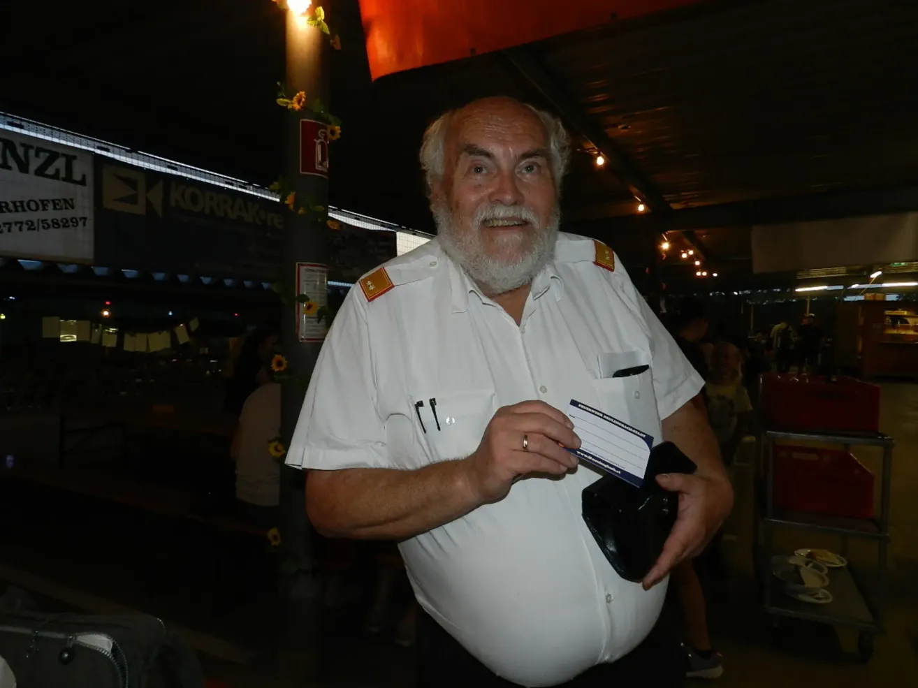Ein älterer Mann mit weißem Bart und Schnurrbart, in einem weißen Uniformhemd gekleidet, hält eine Karte und eine Brieftasche in den Händen. Er steht in einem schwach beleuchteten Bereich mit einem Pfosten und einem Schild hinter ihm.