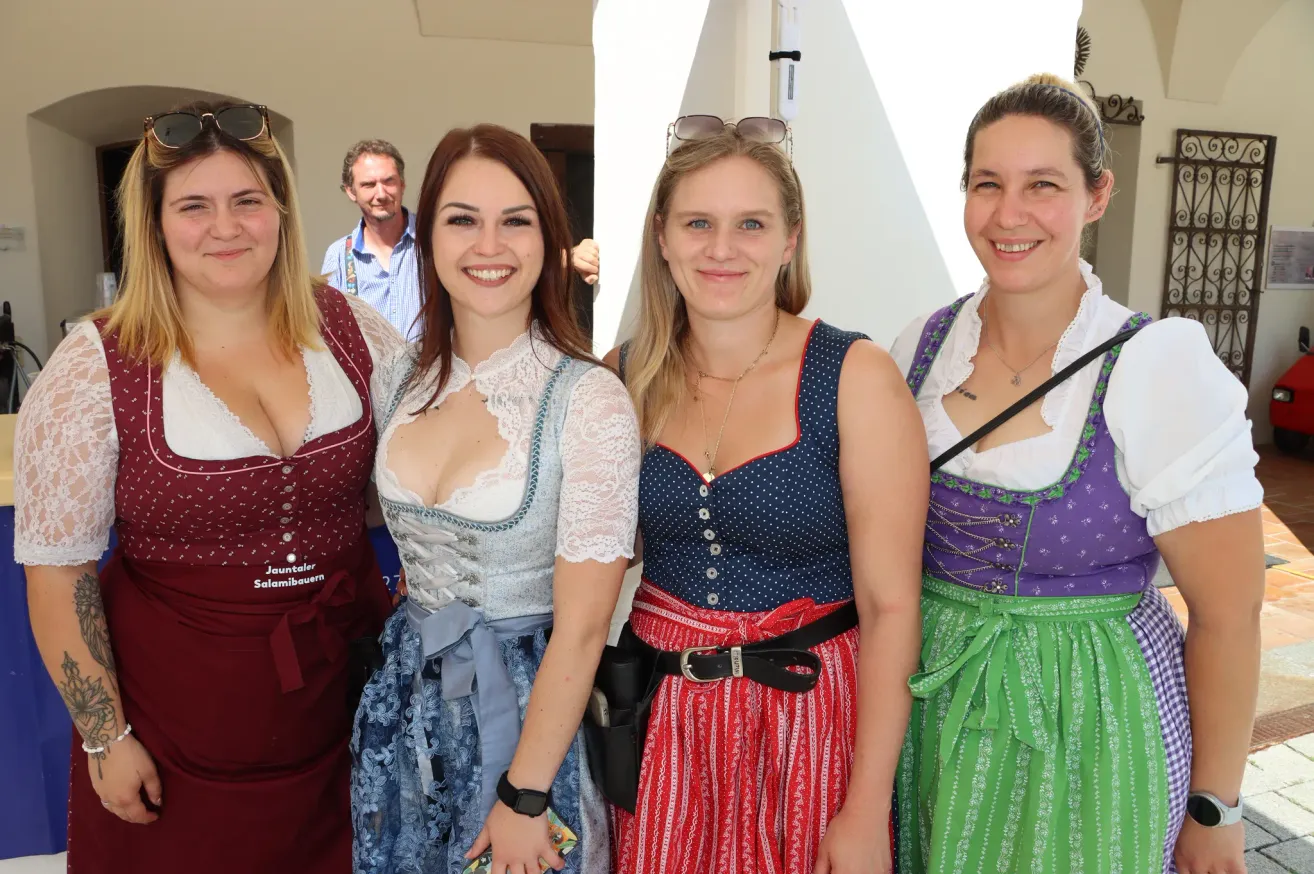 Vier Frauen in traditioneller deutscher Kleidung, die für ein Foto lächeln und posieren. Die Frau auf der linken Seite trägt ein rotes Kleid mit einem weißen Spitzenoberteil, während die Frau in der Mitte ein blaues und weißes gepunktetes Oberteil mit einem roten Rock trägt. Die Frau auf der rechten Seite trägt ein grünes und lila Kleid. Hinter ihnen steht ein Mann in der Nähe einer weißen Säule.