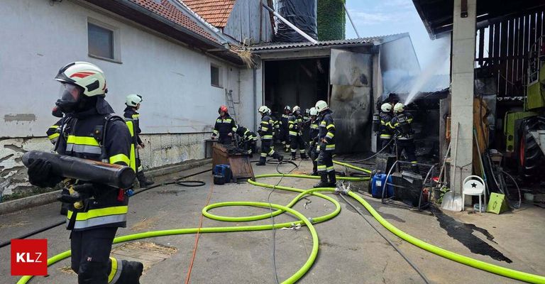 Feuerwehrleute in Schutzkleidung arbeiten daran, ein Feuer in einer Scheune zu löschen. Rauch ist sichtbar, und sie verwenden Schläuche, um Wasser zu sprühen.