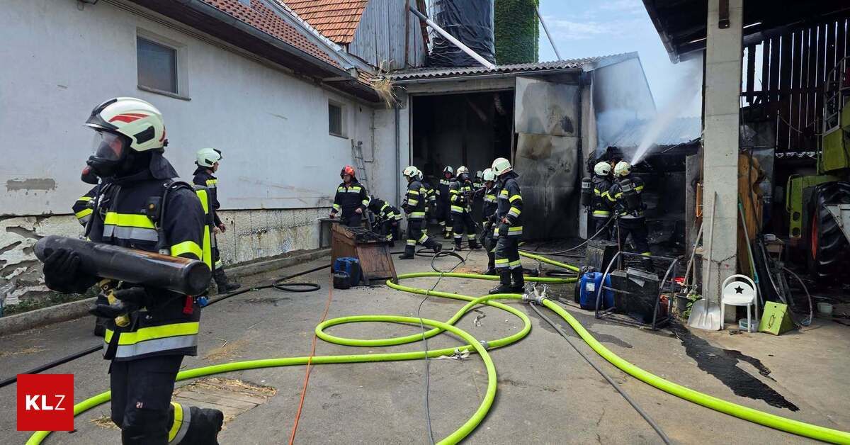 Feuerwehrleute in Schutzkleidung arbeiten daran, ein Feuer in einer Scheune zu löschen. Rauch ist sichtbar, und sie verwenden Schläuche, um Wasser zu sprühen.
