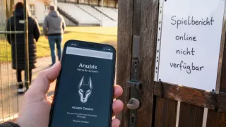 Eine Person hält ein Smartphone mit dem Anubis-Sicherheitssystem-Logo und einer Zugriff verweigert-Meldung. Eine Person geht im Hintergrund in der Nähe eines Holztors.